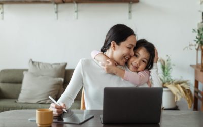 Woman working remotely at home on laptop receiving hug from daughter